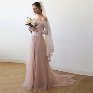 Puede incluir: Un vestido de novia de tul rosa claro con un corpi&ntilde;o de encaje de hombros descubiertos y mangas largas. El vestido tiene una falda fluida y una cola. La novia lleva un ramo de flores blancas y melocot&oacute;n.