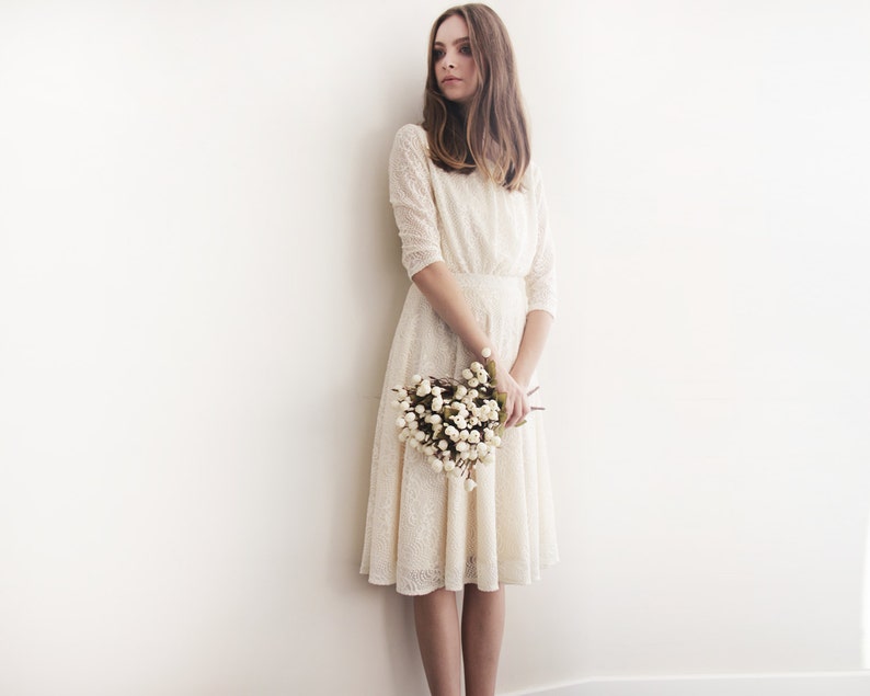 May include: A woman in a white lace dress holds a bouquet of white flowers. She is standing in front of a white wall.