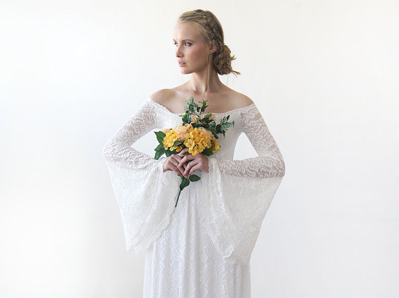 May include: A white off-the-shoulder lace wedding dress with long bell sleeves. The dress has a fitted bodice and a flowing skirt. The model is holding a bouquet of yellow flowers.