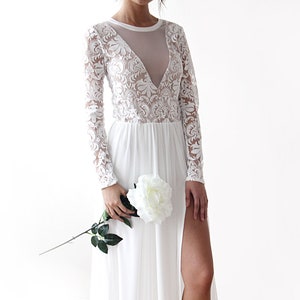 Puede incluir: Un vestido maxi blanco con mangas largas y una superposici&oacute;n de encaje. El vestido tiene un escote pronunciado y una abertura en la pierna hasta el muslo. La modelo sostiene una rosa blanca.