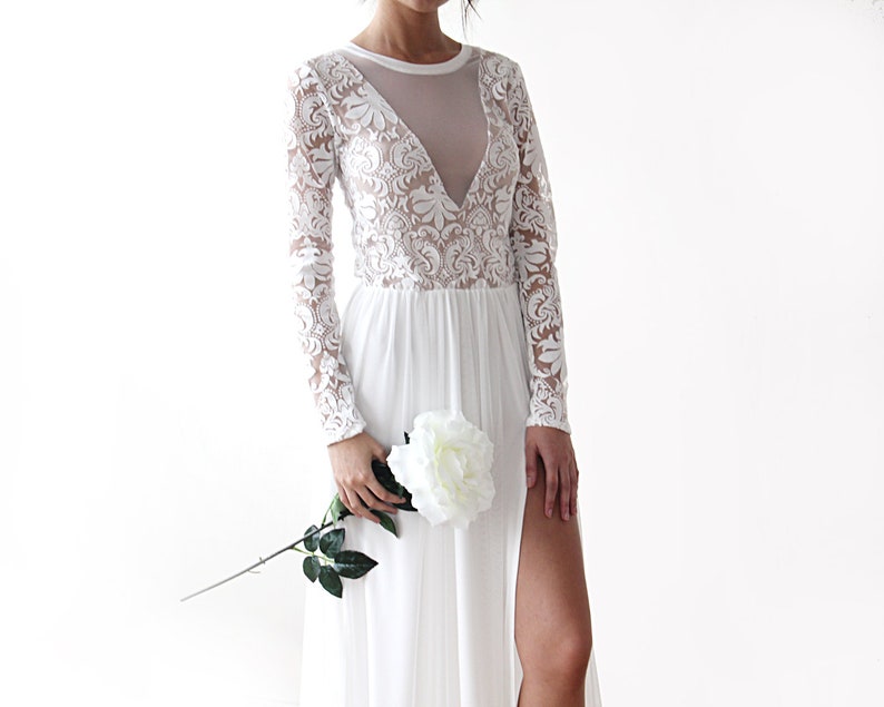 Puede incluir: Un vestido maxi blanco con mangas largas y una superposici&oacute;n de encaje. El vestido tiene un escote pronunciado y una abertura en la pierna hasta el muslo. La modelo sostiene una rosa blanca.