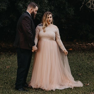 Puede incluir: Una pareja posa para un retrato de boda. La mujer lleva un vestido de novia de tul rosa claro con detalles de encaje. El hombre lleva un traje negro y corbata.
