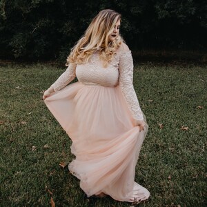 Puede incluir: Una mujer con un vestido de tul rosa claro con mangas de encaje. El vestido tiene un corpi&ntilde;o ajustado y una falda fluida. Est&aacute; de pie en un campo de hierba.