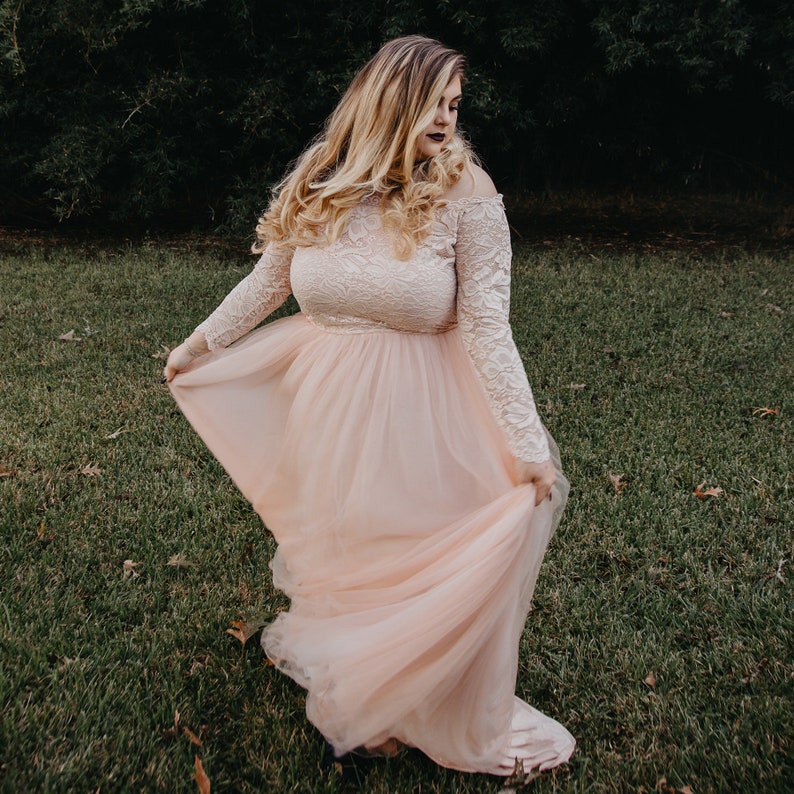 Puede incluir: Una mujer con un vestido de tul rosa claro con mangas de encaje. El vestido tiene un corpi&ntilde;o ajustado y una falda fluida. Est&aacute; de pie en un campo de hierba.