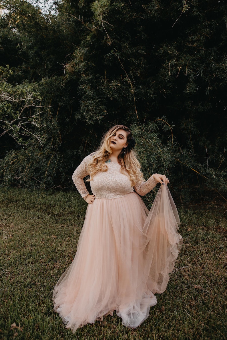 Puede incluir: Una mujer con un vestido de tul rosa claro con detalles de encaje y mangas largas. Est&aacute; de pie en un &aacute;rea cubierta de hierba con un fondo verde oscuro.