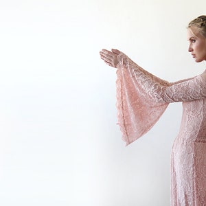 May include: A woman wearing a long, off-the-shoulder, blush pink lace dress with bell sleeves. The dress has a fitted bodice and a flowing skirt.