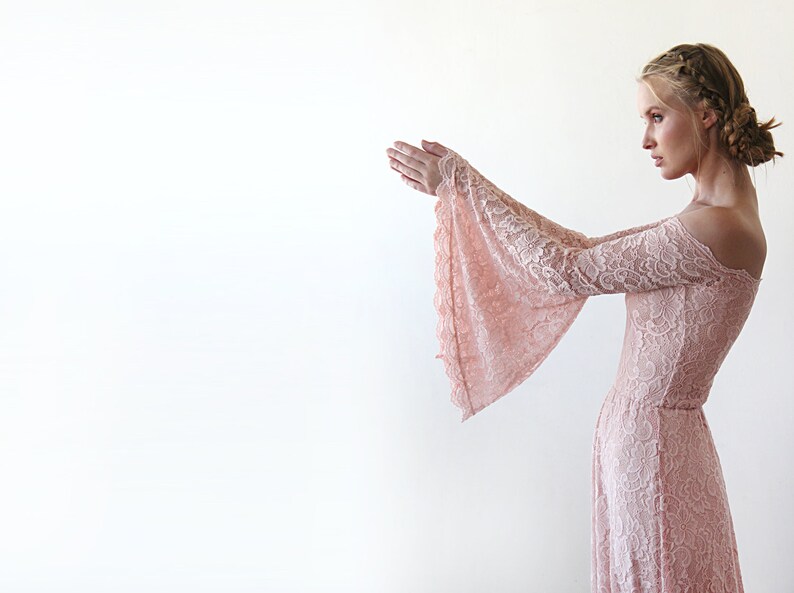 May include: A woman wearing a long, off-the-shoulder, blush pink lace dress with bell sleeves. The dress has a fitted bodice and a flowing skirt.