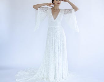 May include: A white lace wedding dress with a deep V-neckline and long, flowing sleeves. The dress has a fitted bodice and a flowing skirt.