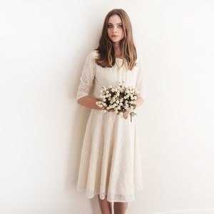 May include: A woman in a white lace dress holds a bouquet of white flowers. She is standing in front of a white wall.