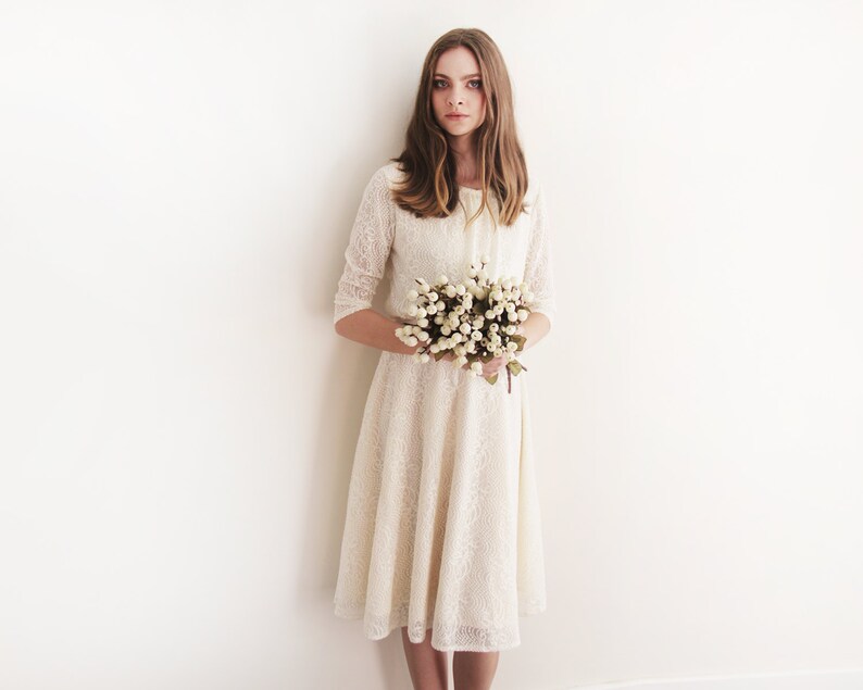 May include: A woman in a white lace dress holds a bouquet of white flowers. She is standing in front of a white wall.