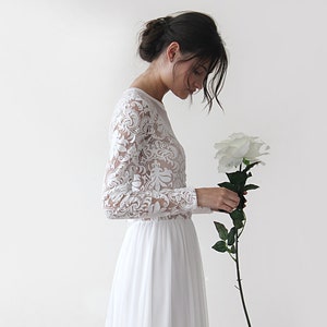 Puede incluir: Un vestido de novia de encaje blanco con mangas largas y una falda fluida. El vestido se combina con una sola rosa blanca.