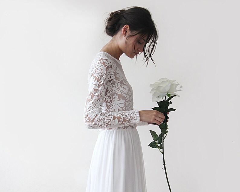 Puede incluir: Un vestido de novia de encaje blanco con mangas largas y una falda fluida. El vestido se combina con una sola rosa blanca.