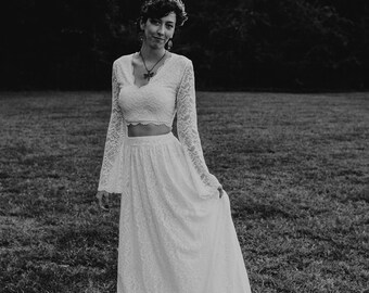 May include: A woman wearing a white lace two-piece bridal outfit with a long skirt and a cropped top. She is standing in a grassy field with trees in the background.