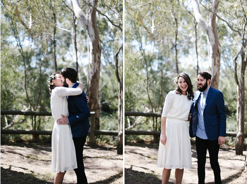 May include: A couple in a forest setting, the bride is wearing a white lace dress and a floral crown, the groom is wearing a blue suit jacket and a light blue shirt. The couple is kissing and smiling.