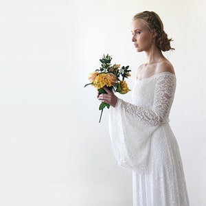 May include: A white off-the-shoulder lace wedding dress with long bell sleeves. The dress is flowing and has a romantic feel. The model is holding a bouquet of yellow and orange flowers.