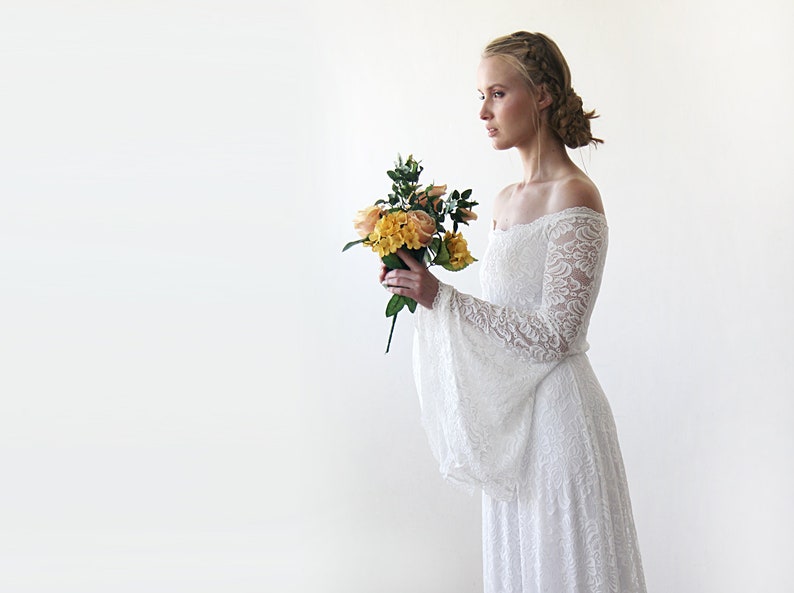 May include: A white off-the-shoulder lace wedding dress with long bell sleeves. The dress is flowing and has a romantic feel. The model is holding a bouquet of yellow and orange flowers.