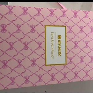 Op de afbeelding: Een rechthoekige roze kartonnen doos met een herhalend patroon van roze linten gestrikt in strikjes over het oppervlak. Een centraal wit rechthoekig label met een gouden rand toont de tekst STANLEY en LOVESHACKFANCY in gouden letters.