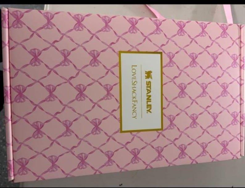 Op de afbeelding: Een rechthoekige roze kartonnen doos met een herhalend patroon van roze linten gestrikt in strikjes over het oppervlak. Een centraal wit rechthoekig label met een gouden rand toont de tekst STANLEY en LOVESHACKFANCY in gouden letters.