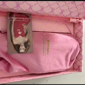 Op de afbeelding: Een roze doos met strikpatroon bevat een roze stoffen zakje met de in goud gedrukte logo's van Stanley en LoveShackFancy. Een rechthoekige kaart toont een persoon in een rode jurk met de tekst Welcome to the world of LoveShackFancy.
