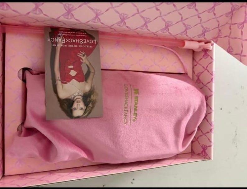 Op de afbeelding: Een roze doos met strikpatroon bevat een roze stoffen zakje met de in goud gedrukte logo's van Stanley en LoveShackFancy. Een rechthoekige kaart toont een persoon in een rode jurk met de tekst Welcome to the world of LoveShackFancy.