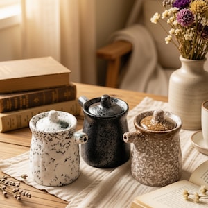 Può includere: Tre versatori in ceramica con coperchio, ognuno caratterizzato da una finitura maculata. Un versatore è bianco con macchie nere, uno è nero con superficie testurizzata e uno è color cuoio con macchie bianche. Ognuno ha un piccolo beccuccio laterale.