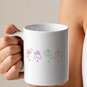 May include: A white ceramic mug featuring a colourful graphic of four hand signs representing the British Sign Language alphabet, spelling out the name KATE in red, purple, green, and orange, with each letter positioned below its corresponding hand sign.