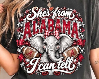 May include: A charcoal gray short-sleeve t-shirt featuring a graphic print of an elephant wearing a red and white plaid bow. The text reads She's from ALABAMA I can tell, surrounded by red bows, footballs, and stars.
