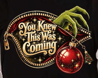 May include: A black t-shirt featuring a graphic of a green, clawed hand unzipping a circular opening. The text reads "You Knew This Was Coming" in gold lettering. A shiny red Christmas ornament hangs from the zipper pull, accented by a small tree.