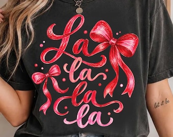 May include: A charcoal gray short-sleeve t-shirt featuring a graphic print of red ribbons and the text "La la la" in a cursive font. The design includes small red dots scattered around the ribbons and text on the front of the shirt.