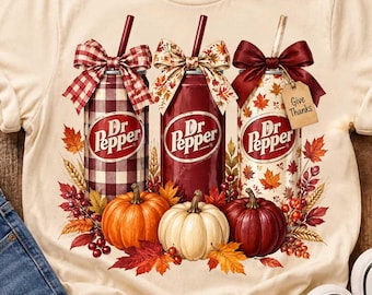 May include: A cream-colored t-shirt featuring a graphic of three Dr Pepper cans topped with bows and straws, surrounded by autumn leaves and pumpkins. The cans are decorated with plaid, solid red, and leaf patterns, with a tag reading Give Thanks.