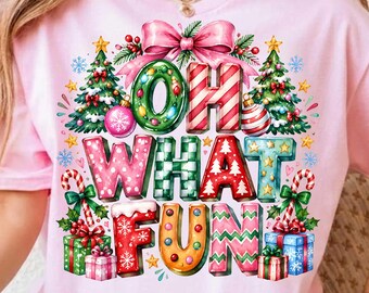 May include: A light pink short-sleeve t-shirt featuring a colorful graphic print with the text "OH WHAT FUN". The design includes decorated Christmas trees, candy canes, gift boxes, a large pink bow, and various holiday-themed patterns.