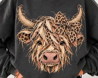 May include: A charcoal gray crewneck sweatshirt featuring a graphic of a Highland cow with a textured, frayed appearance. The cow has a leopard print bow on its head and leopard print accents on its ears, set against the dark fabric.