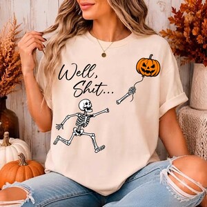 Puede incluir: Una camiseta de color crema con un gr&aacute;fico negro de un esqueleto corriendo mientras sostiene una cuerda atada a una calabaza naranja. El texto "Well, Shit..." est&aacute; impreso en letra cursiva negra sobre la ilustraci&oacute;n del esqueleto.
