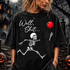Puede incluir: Una camiseta negra con efecto lavado al &aacute;cido que presenta un gr&aacute;fico blanco de un esqueleto corriendo, un globo rojo y el texto "Well, Shit..." en letra cursiva. La prenda tiene cuello redondo, mangas cortas y una textura desgastada.