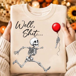 Puede incluir: Una sudadera de cuello redondo en color beige con un gr&aacute;fico negro de un esqueleto corriendo que sostiene un globo rojo. El texto "Well, Shit..." est&aacute; impreso en letra cursiva negra sobre el esqueleto en la parte frontal de la prenda.