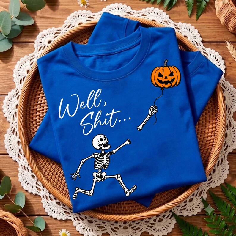 Puede incluir: Una camiseta azul real con un gr&aacute;fico blanco de un esqueleto corriendo hacia un globo naranja de calabaza flotante. El texto "Well, Shit..." est&aacute; impreso en letra cursiva blanca sobre el esqueleto en la parte frontal de la prenda.