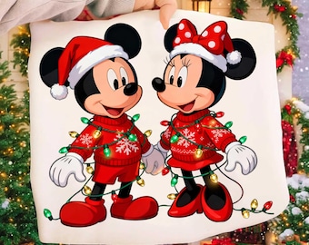 May include: A white fabric item featuring a graphic print of Mickey and Minnie Mouse wearing red Santa hats and matching red sweaters with white snowflake patterns. Both characters are wrapped in a string of colorful Christmas lights.