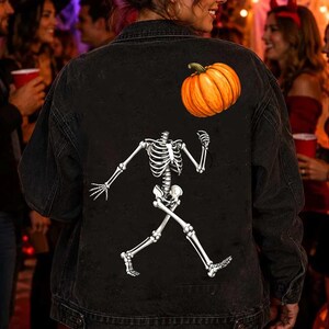 Puede incluir: Una chaqueta de mezclilla negra con un estampado gr&aacute;fico en la parte posterior. El dise&ntilde;o muestra un esqueleto blanco en pose de caminar con una calabaza naranja colocada sobre el &aacute;rea del hombro del esqueleto. La chaqueta tiene mangas largas.