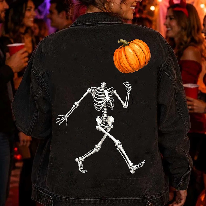 Puede incluir: Una chaqueta de mezclilla negra con un estampado gr&aacute;fico en la parte posterior. El dise&ntilde;o muestra un esqueleto blanco en pose de caminar con una calabaza naranja colocada sobre el &aacute;rea del hombro del esqueleto. La chaqueta tiene mangas largas.