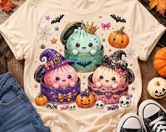 May include: A cream-colored t-shirt featuring a graphic of three cute, round, colorful characters in containers. One is light blue with a gold crown, one is pink with a purple witch hat, and one is light pink with a black witch hat. Includes pumpkins, bats, and skulls.