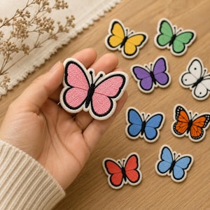 Könnte beinhalten: Eine Sammlung von gestickten Schmetterlingsaufnähern in verschiedenen Farben, darunter Pink, Gelb, Grün, Lila, Weiß, Blau, Orange und Rot. Jeder Aufnäher hat einen schwarz gestickten Körper und Fühler sowie einen weißen Rand um die Flügel.