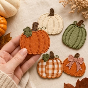 May include: A collection of five embroidered pumpkin patches featuring various designs. One is bright orange with a green leaf, one is cream, one is sage green, one has an orange and white gingham pattern, and one is orange with a pink bow.