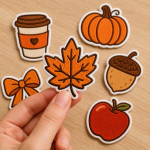 Peut inclure: Un ensemble de six écussons brodés aux motifs automnaux comprenant une tasse de café avec un cœur, une citrouille, une feuille d'érable, un nœud, un gland et une pomme rouge, tous avec des bordures blanches et des coutures orange, marron et rouge.