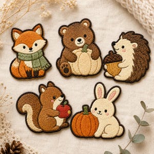 May include: Five embroidered patches featuring woodland animals: a fox in a green scarf, a brown bear holding a pumpkin, a hedgehog with an acorn, a squirrel with a red apple, and a cream-colored rabbit sitting next to an orange pumpkin.