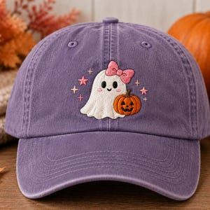 May include: A purple baseball cap featuring an embroidered design of a white ghost with a pink bow, holding an orange jack-o'-lantern. The ghost is surrounded by small pink and yellow star-shaped accents, centered on the front panel of the cap.