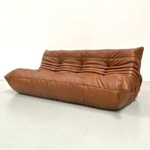 May include: A low-profile, three-seater sofa upholstered in cognac-colored leather. The piece features a distinctively tufted, pleated design across the backrest and seat, creating a soft, rounded silhouette with no visible legs or frame structure.