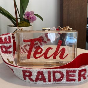 May include: A clear rectangular acrylic box clutch with a gold-tone metal frame and clasp. The front features the word Tech in red script. It includes a white beaded shoulder strap with the word RAIDERS in red beads and gold star accents.