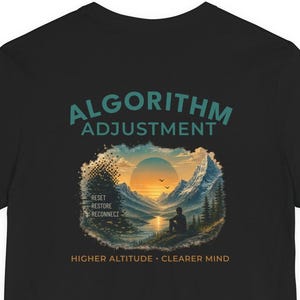 May include: A black t-shirt featuring a graphic of a person sitting by a lake looking at mountains during sunset. The text reads ALGORITHM ADJUSTMENT, RESET RESTORE RECONNECT, and HIGHER ALTITUDE CLEARER MIND in teal and orange lettering.