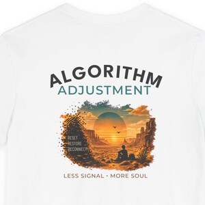 May include: A white t-shirt featuring a graphic print with the text ALGORITHM ADJUSTMENT at the top. Below, a desert landscape illustration shows a sunset, a person sitting, and the words RESET RESTORE RECONNECT. The text LESS SIGNAL MORE SOUL appears at the bottom.