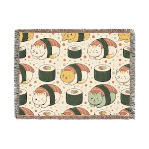Op de afbeelding: Een geweven plaid met een herhalend patroon van cartoonkatten verkleed als sushibroodjes, afgewisseld met maki-rollen en kleine kleurrijke stippen op een crèmekleurige achtergrond. De deken heeft een textuurrijke franje aan alle vier de zijden.